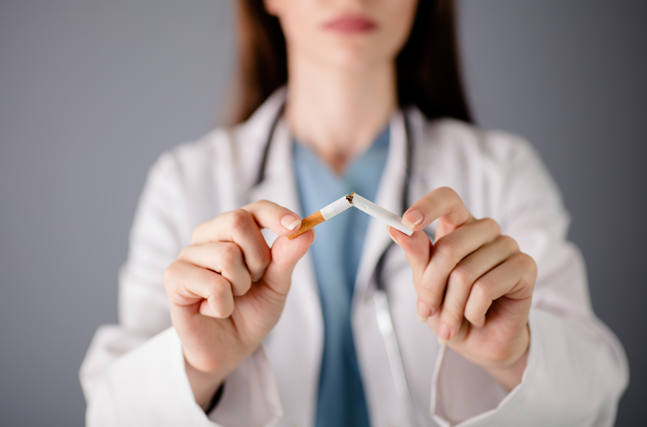 Cigarette snapped in half in the foreground by a person wearing a medical coat and stethoscope blurred in the background.